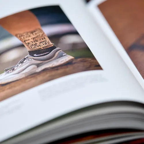 Close-up of an open photo book showing a picture of an athlete's lower legs wearing running shoes and socks with motivational text, standing on a dirt surface.