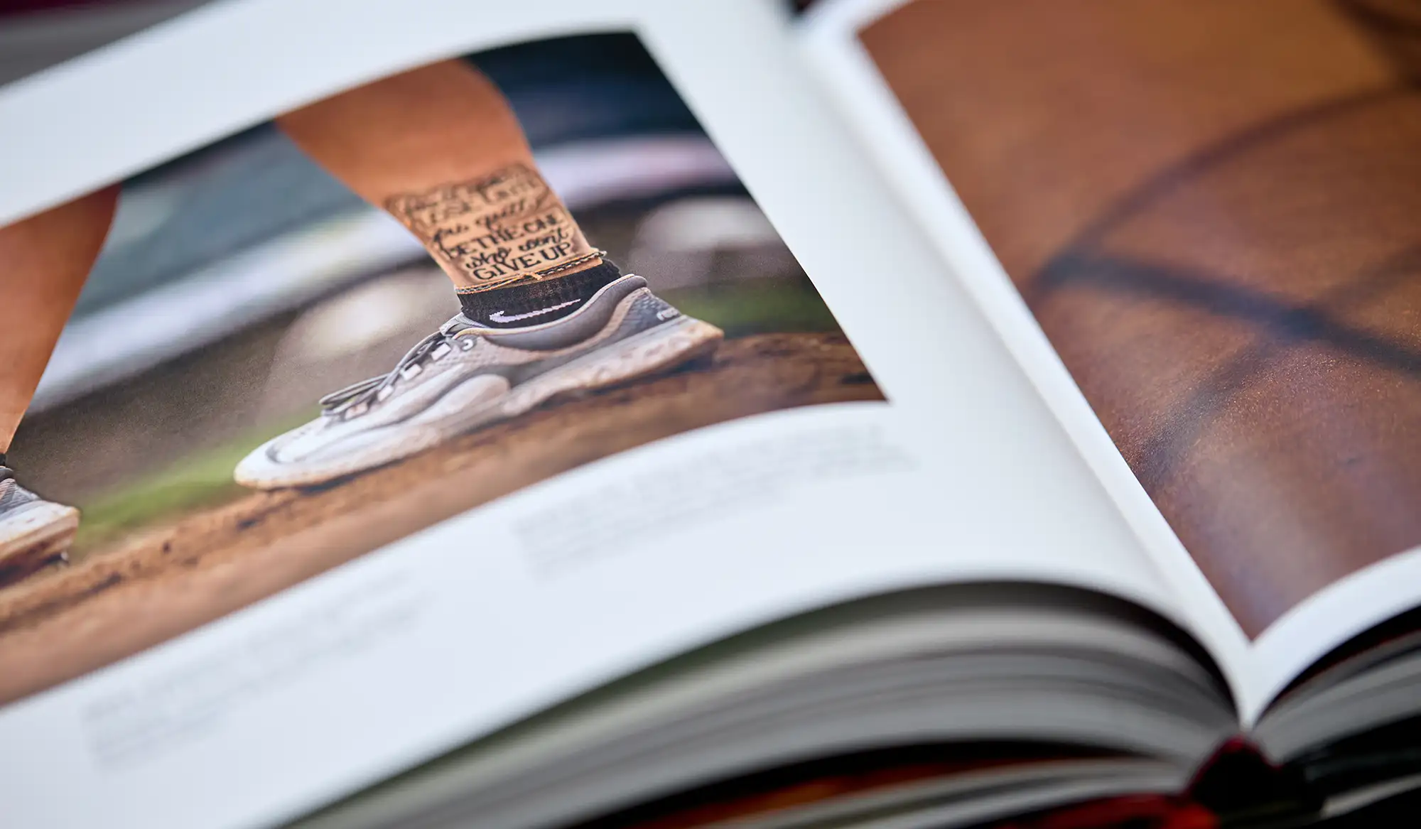 Close-up of an open photo book showing a picture of an athlete's lower legs wearing running shoes and socks with motivational text, standing on a dirt surface.