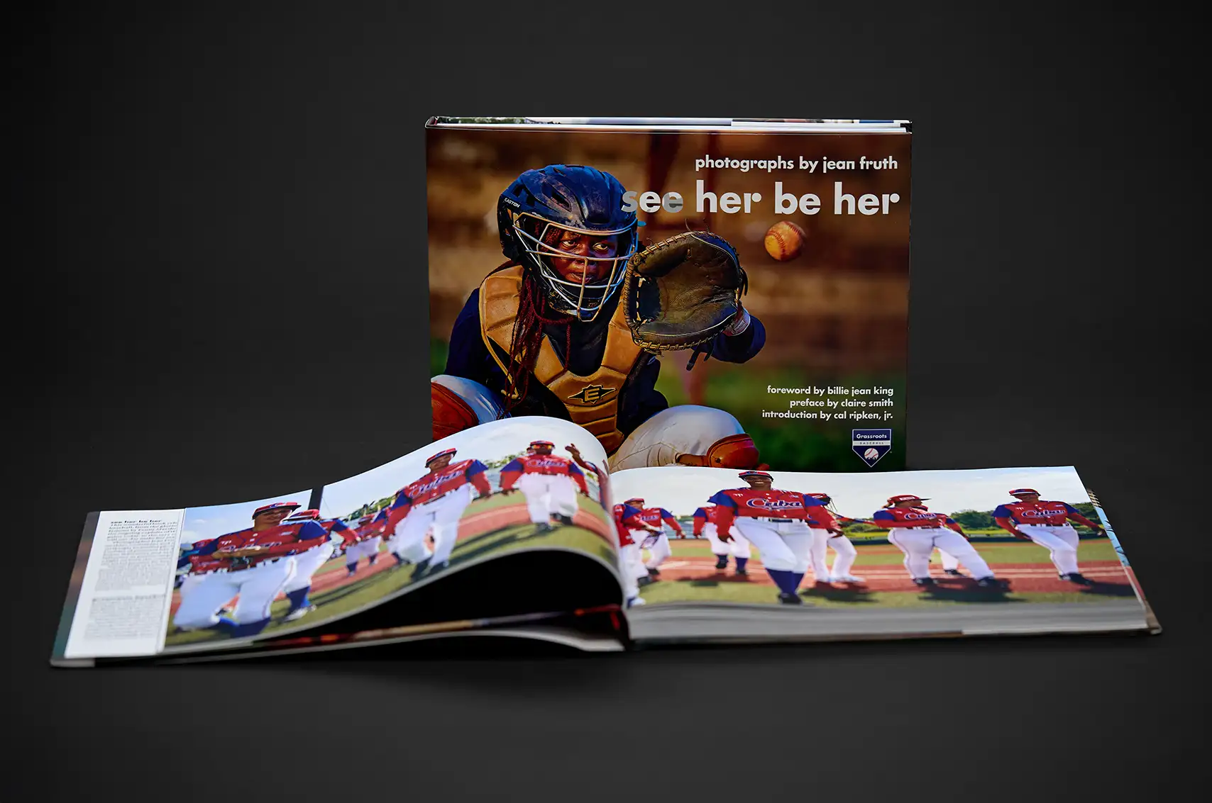 Hardcover book titled 'See Her Be Her' by Jean Fruth, featuring a female baseball catcher on the cover, displayed with the book open showing a colorful photo of a women's baseball team walking on the field.