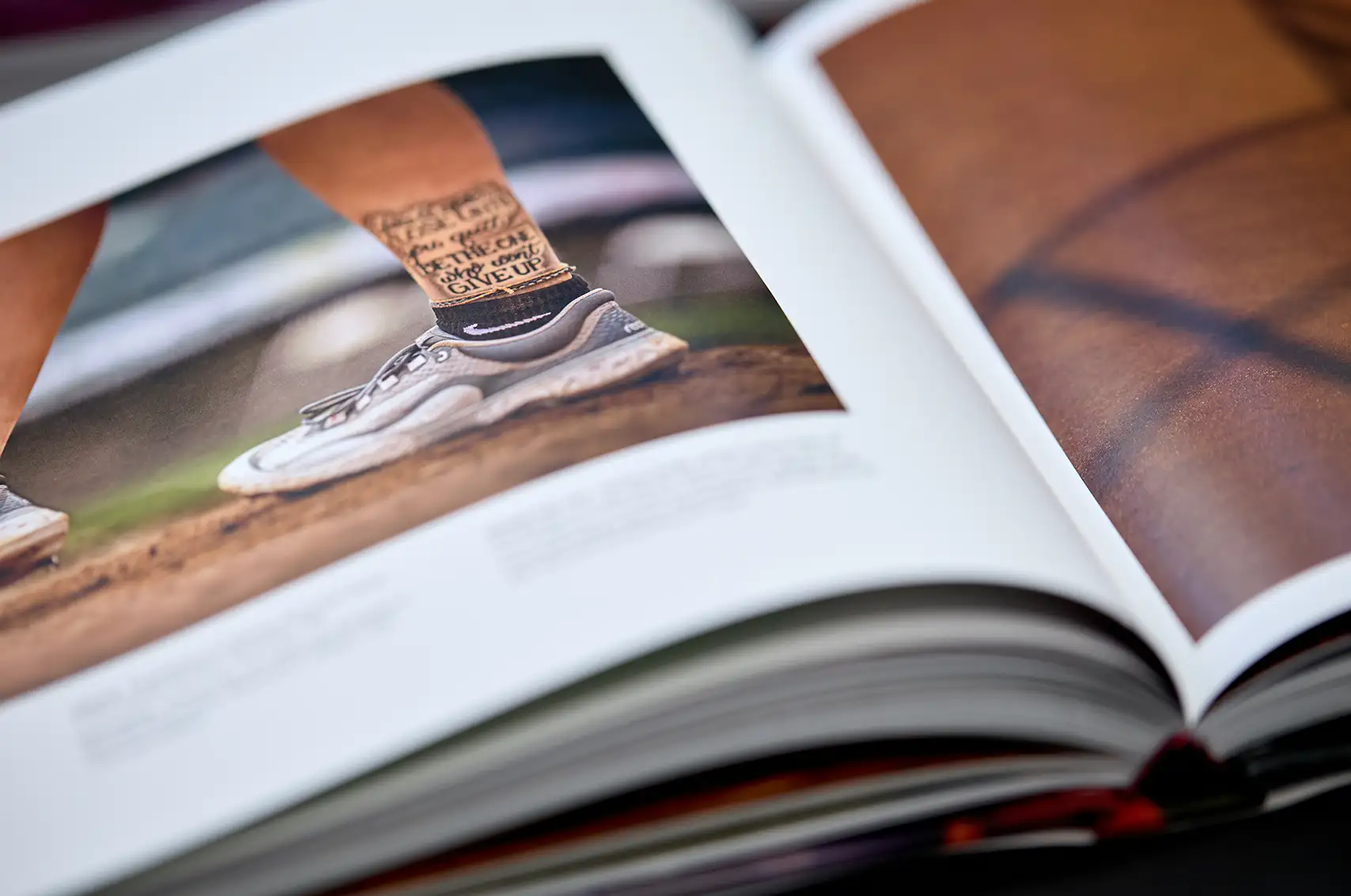Close-up of an open photo book showing a picture of an athlete's lower legs wearing running shoes and socks with motivational text, standing on a dirt surface.